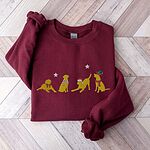 Maroon crewneck sweatshirt laid flat on a chevron wood floor, embroidered with four playful yellow labs in a Santa hat, an elf hat, a red bow and a striped scarf under silver stars
