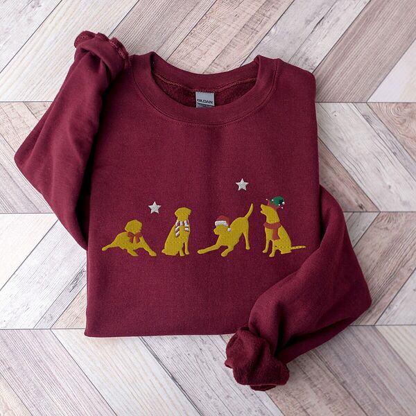 Maroon crewneck sweatshirt laid flat on a chevron wood floor, embroidered with four playful yellow labs in a Santa hat, an elf hat, a red bow and a striped scarf under silver stars