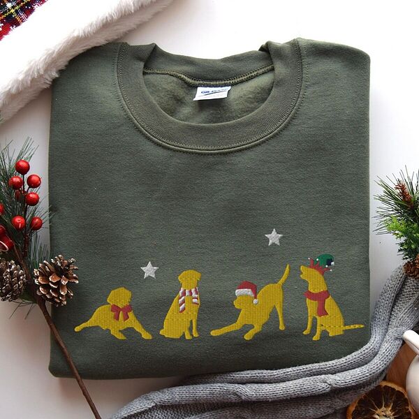 Military Green crewneck sweatshirt laid flat with pine cones and red berries, embroidered with four playful yellow labs in a Santa hat, an elf hat, a red bow and a striped scarf under silver stars