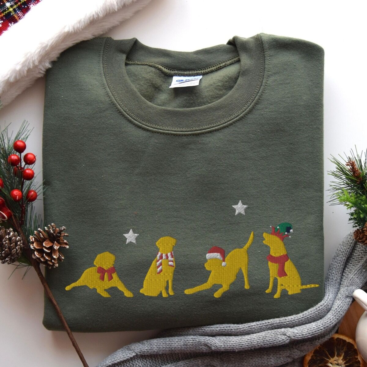 Yellow Lab Christmas Sweatshirt – Military Green Military Green crewneck sweatshirt laid flat with pine cones and red berries, embroidered with four playful yellow labs in a Santa hat, an elf hat, a red bow and a striped scarf under silver stars