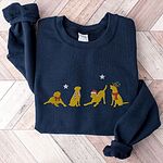 Navy crewneck sweatshirt laid flat on a chevron wood floor, embroidered with four playful yellow labs in a Santa hat, an elf hat, a red bow and a striped scarf under silver stars