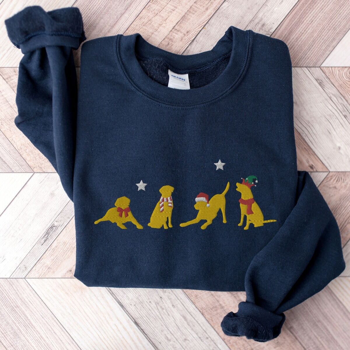 Yellow Lab Christmas Sweatshirt – Navy Navy crewneck sweatshirt laid flat on a chevron wood floor, embroidered with four playful yellow labs in a Santa hat, an elf hat, a red bow and a striped scarf under silver stars