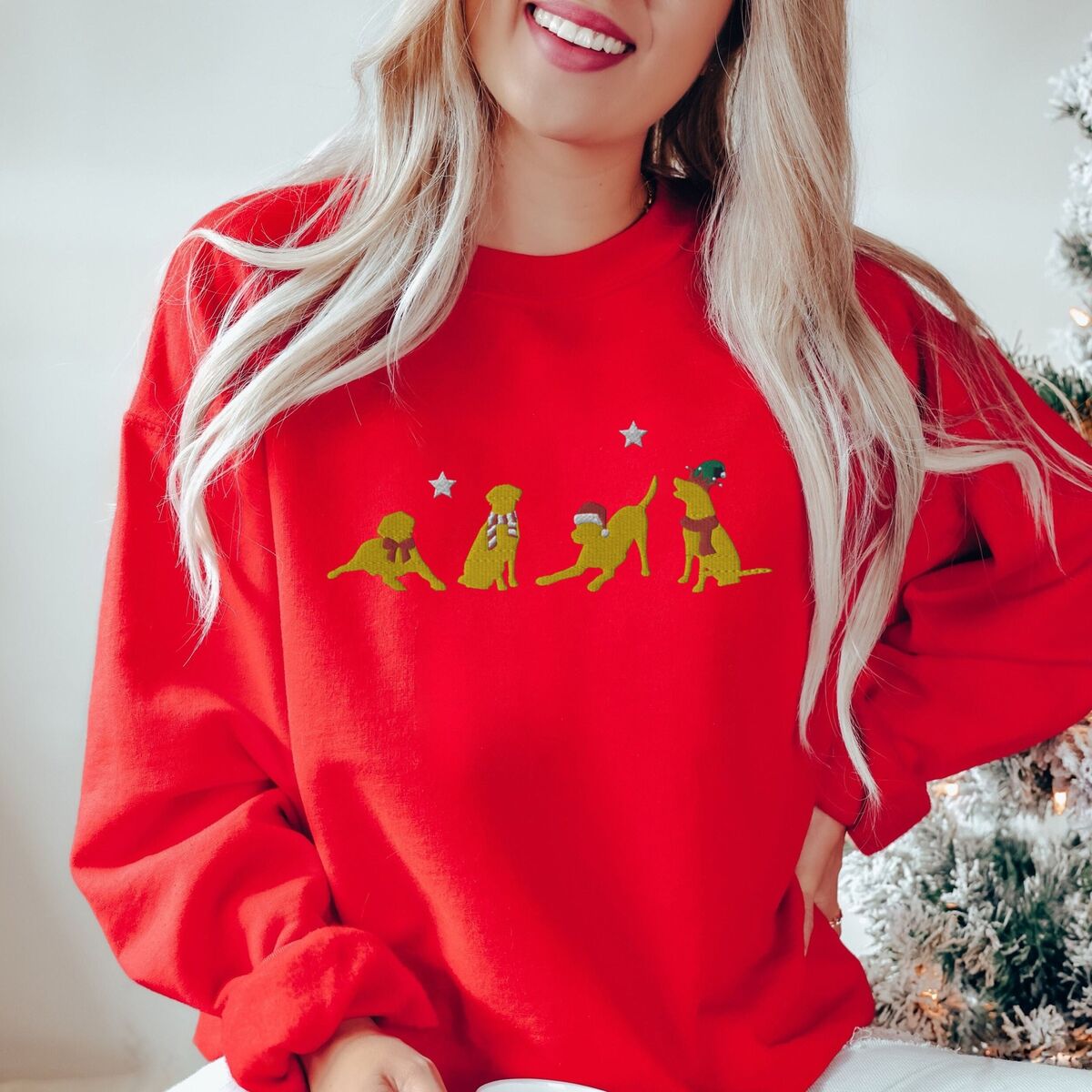 Yellow Lab Christmas Sweatshirt - Red Smiling woman by a Christmas tree in a Red crewneck sweatshirt embroidered with four playful yellow labs in a Santa hat, an elf hat, a red bow and a striped scarf under silver stars