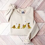 Sand crewneck sweatshirt laid flat on a chevron wood floor, embroidered with four playful yellow labs in a Santa hat, an elf hat, a red bow and a striped scarf under silver stars