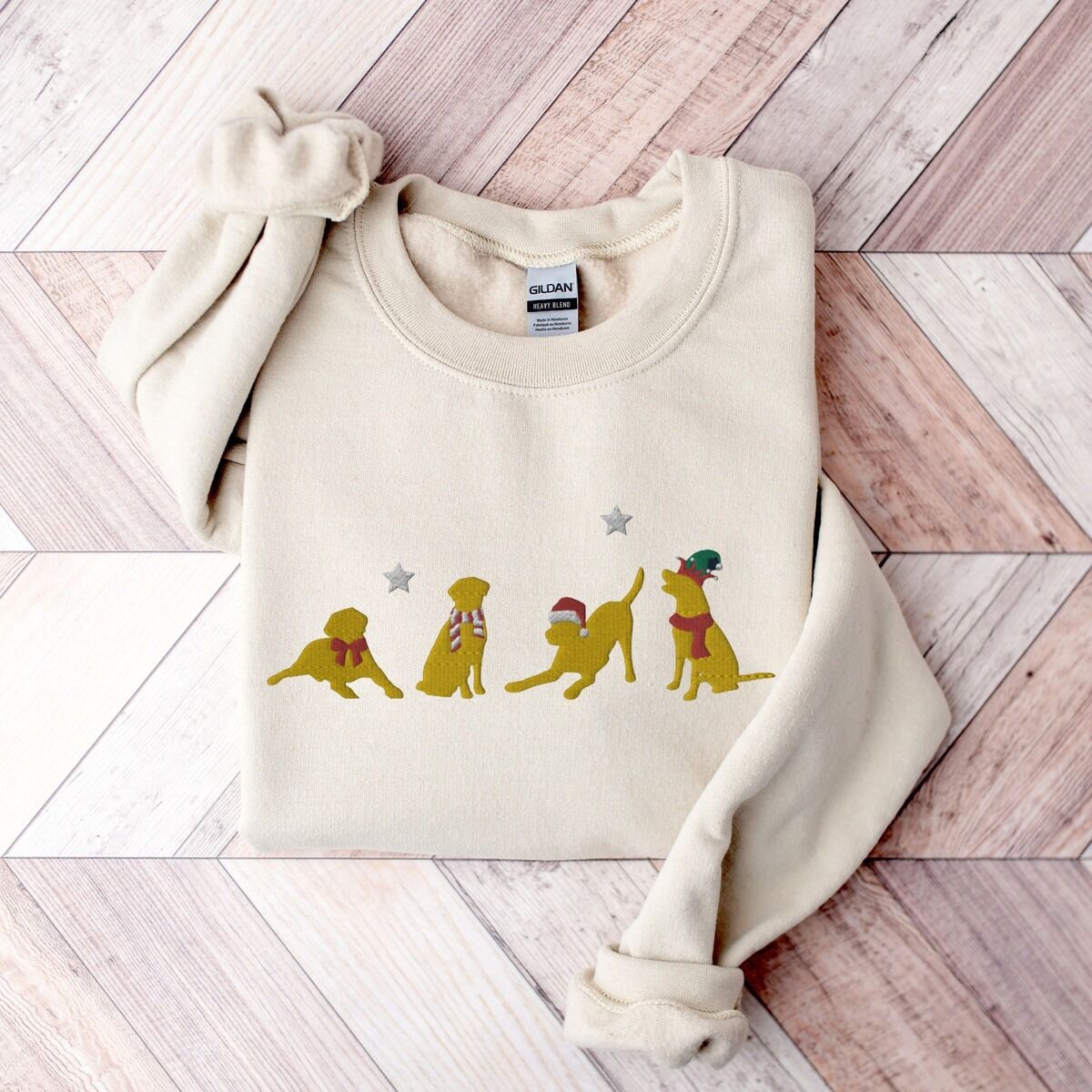 Yellow Lab Christmas Sweatshirt – Sand Sand crewneck sweatshirt laid flat on a chevron wood floor, embroidered with four playful yellow labs in a Santa hat, an elf hat, a red bow and a striped scarf under silver stars