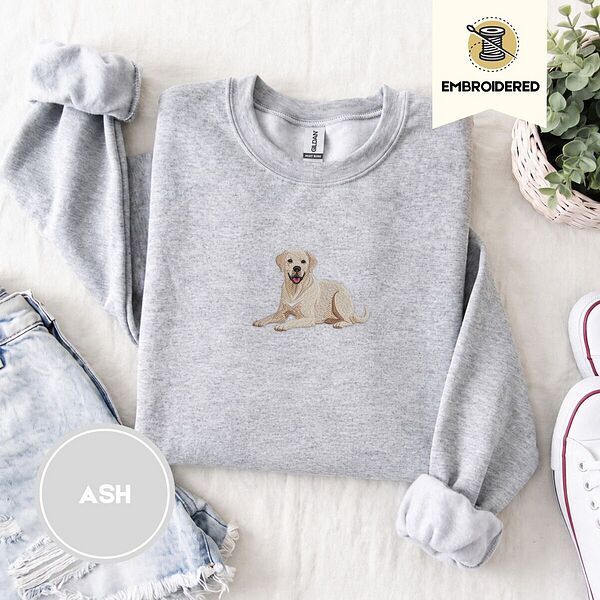 Ash crewneck sweatshirt folded with denim shorts, white sneakers and a potted plant, embroidered with a smiling yellow Labrador lying down with its front paws stretched out