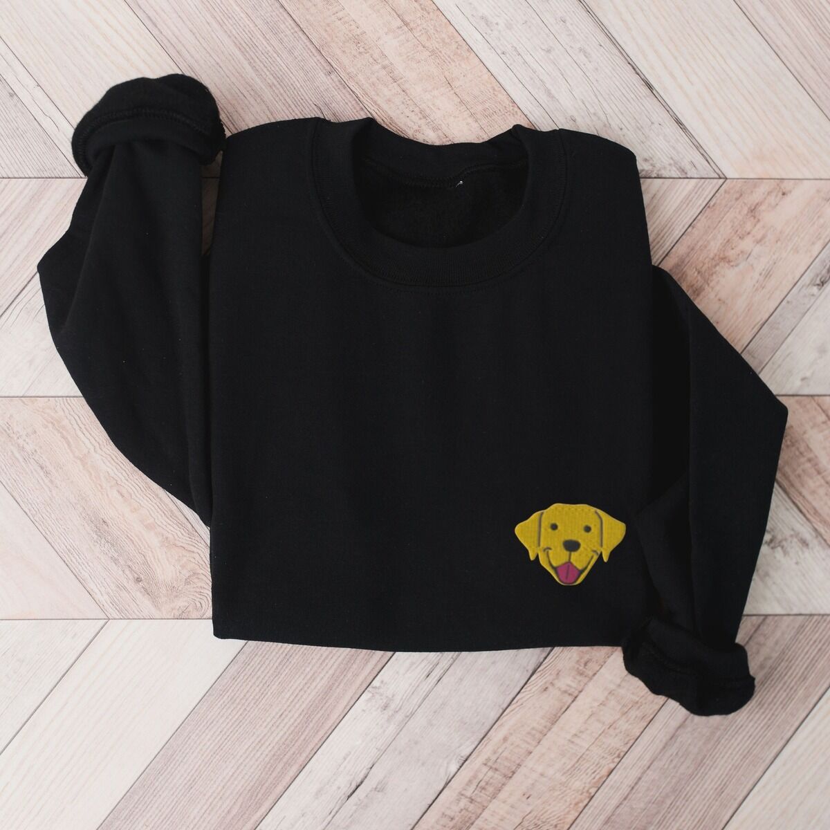 Yellow Lab Face Sweatshirt – Black Black crewneck sweatshirt laid flat on herringbone wood, embroidered with a small smiling yellow Labrador face with floppy ears and a pink tongue on the left chest