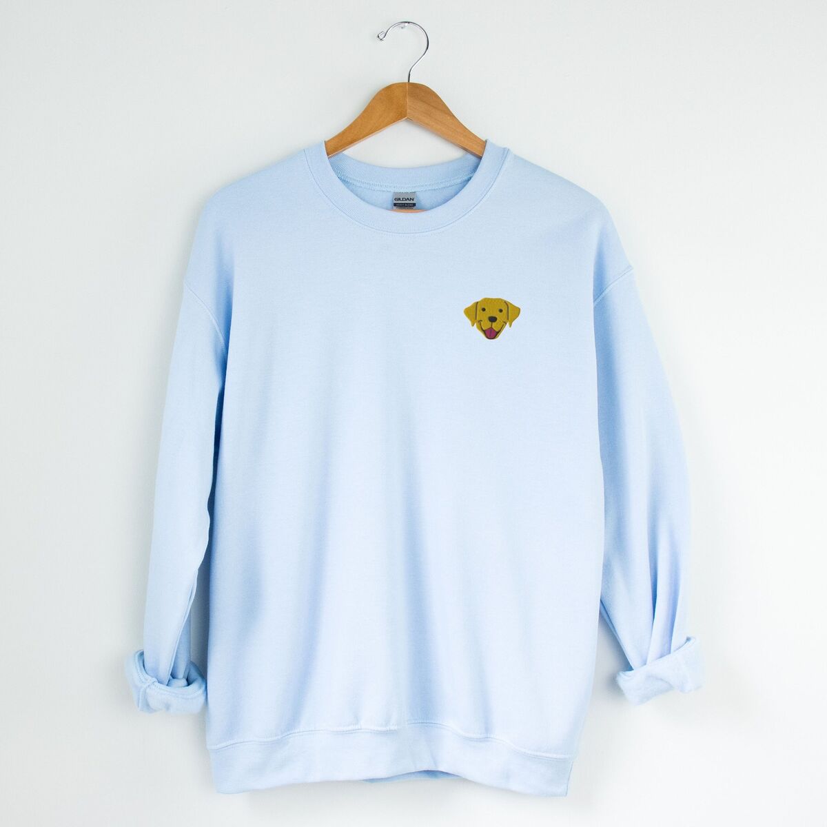 Yellow Lab Face Sweatshirt - Light Blue Light Blue crewneck sweatshirt on a hanger, embroidered with a small smiling yellow Labrador face with floppy ears and a pink tongue on the left chest