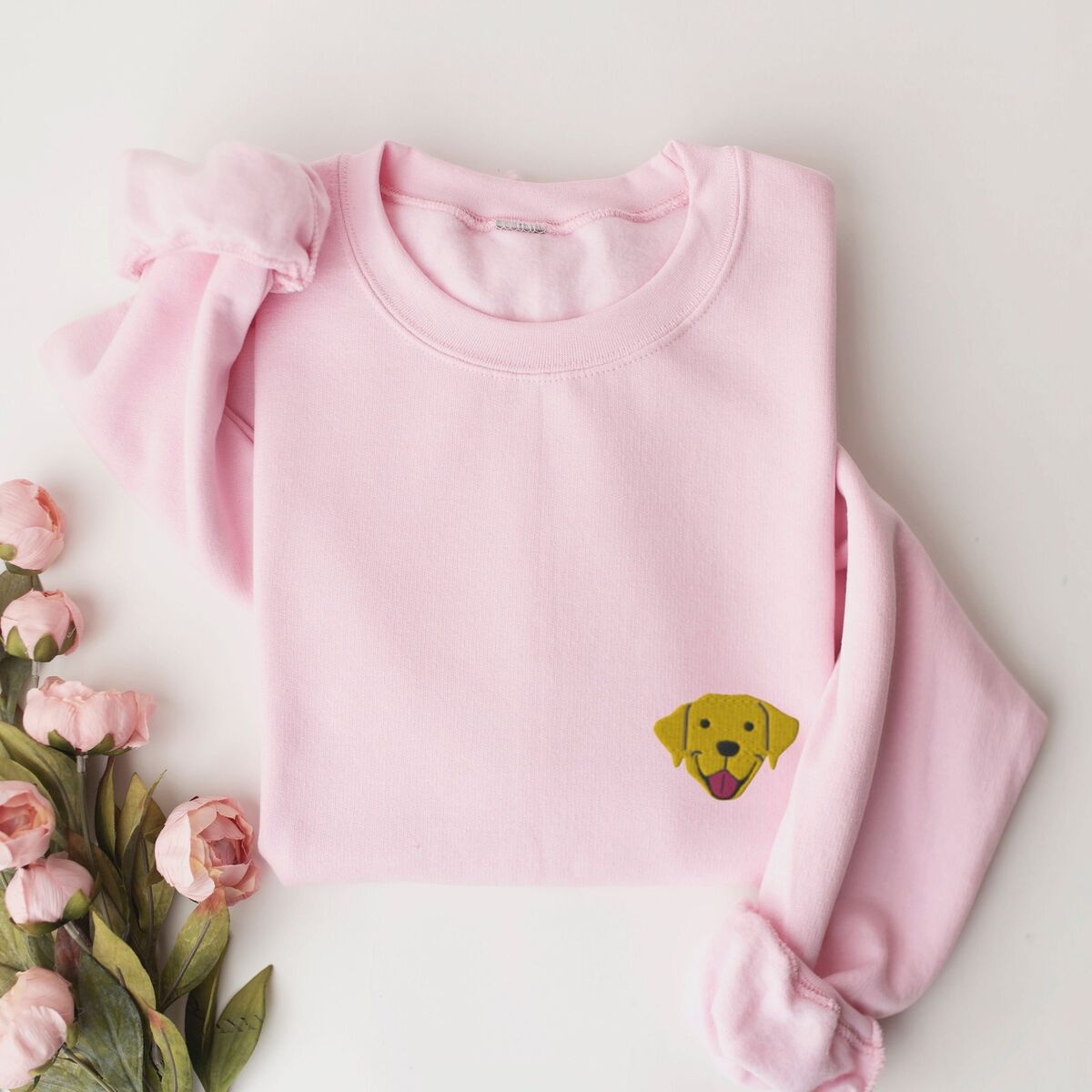 Yellow Lab Face Sweatshirt - Light Pink Light Pink crewneck sweatshirt laid flat with pink roses, embroidered with a small smiling yellow Labrador face with floppy ears and a pink tongue on the left chest