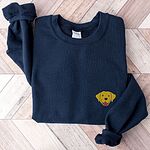 Navy crewneck sweatshirt laid flat on herringbone wood, embroidered with a small smiling yellow Labrador face with floppy ears and a pink tongue on the left chest