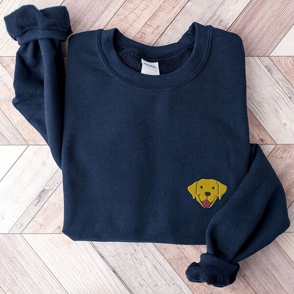 Navy crewneck sweatshirt laid flat on herringbone wood, embroidered with a small smiling yellow Labrador face with floppy ears and a pink tongue on the left chest