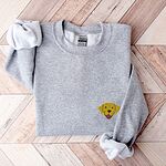Sport Grey crewneck sweatshirt laid flat on herringbone wood, embroidered with a small smiling yellow Labrador face with floppy ears and a pink tongue on the left chest