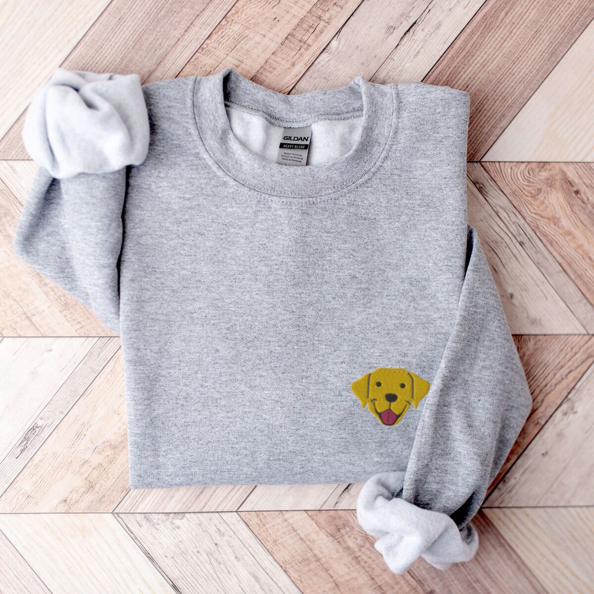 Yellow Lab Face Sweatshirt - Sport Grey Sport Grey crewneck sweatshirt laid flat on herringbone wood, embroidered with a small smiling yellow Labrador face with floppy ears and a pink tongue on the left chest