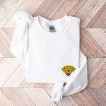 White crewneck sweatshirt laid flat on herringbone wood, embroidered with a small smiling yellow Labrador face with floppy ears and a pink tongue on the left chest