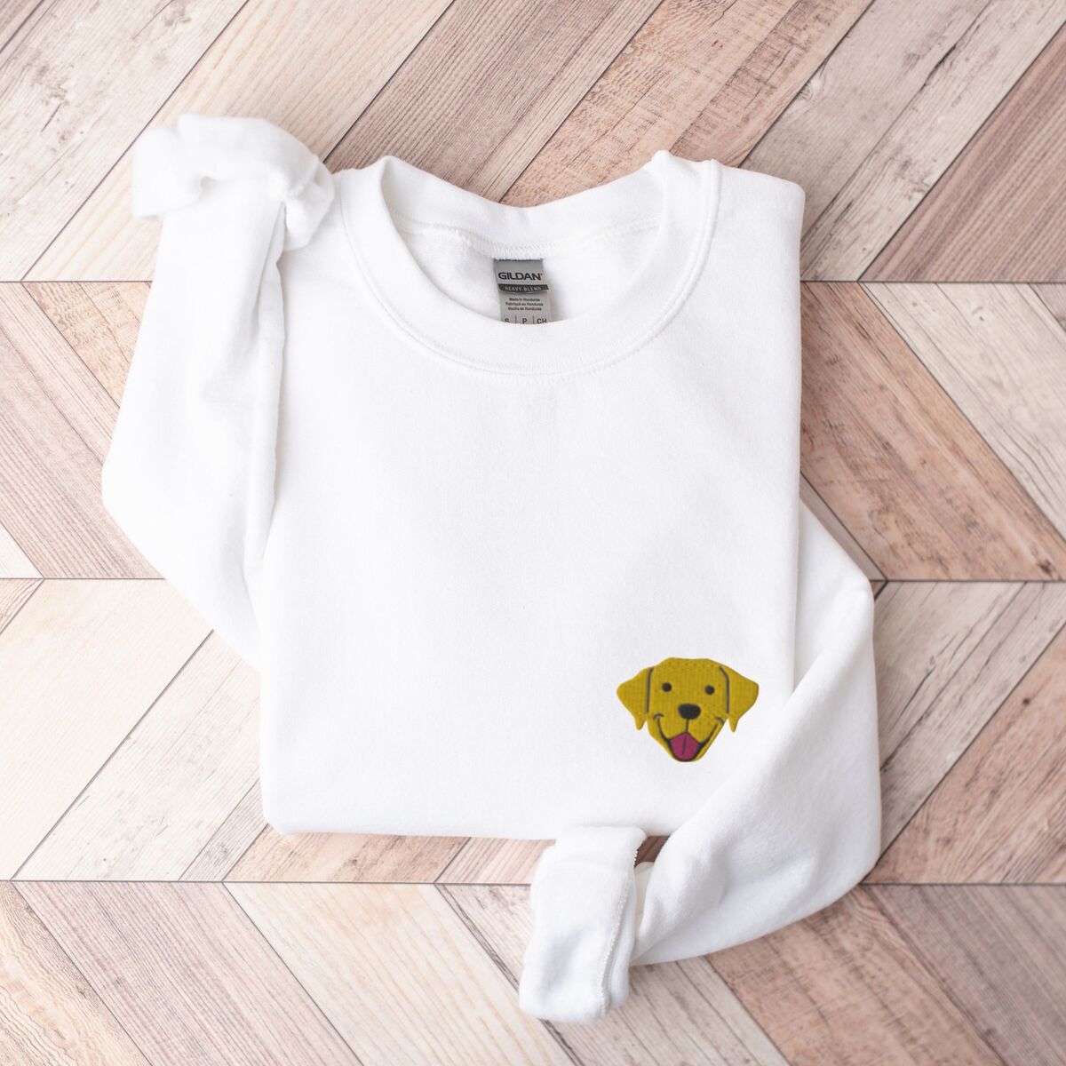 Yellow Lab Face Sweatshirt - White White crewneck sweatshirt laid flat on herringbone wood, embroidered with a small smiling yellow Labrador face with floppy ears and a pink tongue on the left chest
