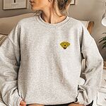 Woman sitting in a light grey crewneck sweatshirt embroidered with a small smiling yellow Labrador face with floppy ears and a pink tongue on the left chest