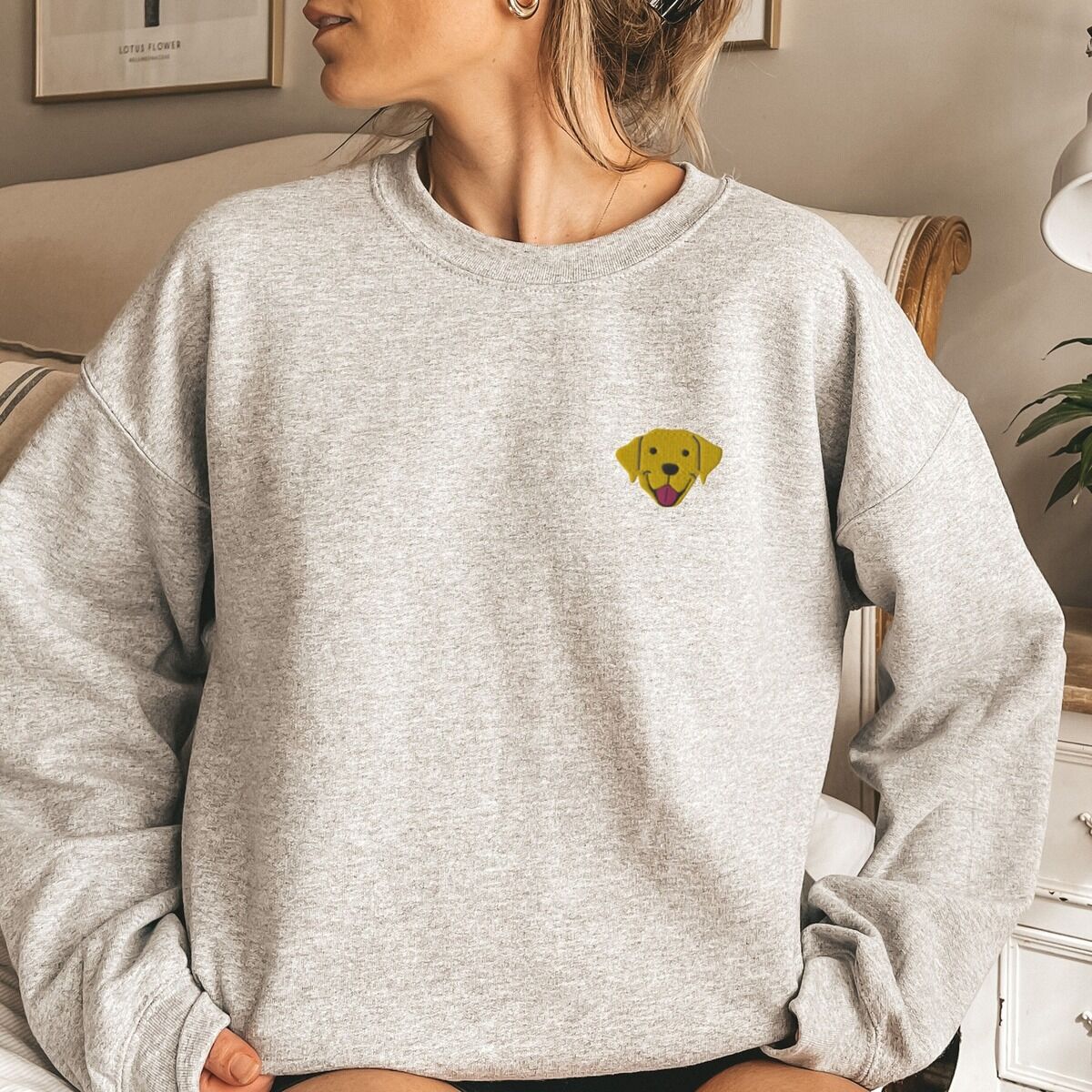 Yellow Lab Face Sweatshirt - worn in grey Woman sitting in a light grey crewneck sweatshirt embroidered with a small smiling yellow Labrador face with floppy ears and a pink tongue on the left chest