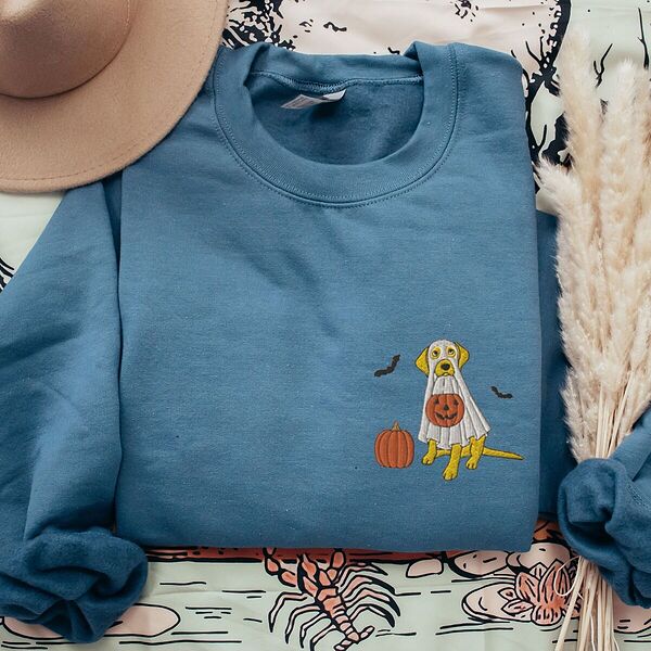 Yellow lab ghost sweatshirt in Indigo Blue laid flat with a felt hat and dried pampas grass, embroidered with a yellow Labrador in a white ghost sheet holding a jack-o'-lantern bucket beside a pumpkin and two bats