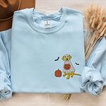 Light Blue crewneck sweatshirt laid flat with dried wheat, embroidered with a yellow Labrador in a ghost sheet with a pumpkin and two bats on the left chest