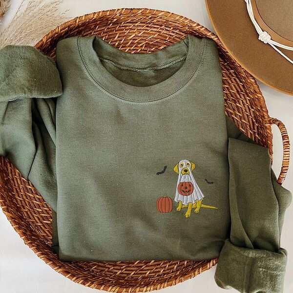 Military Green crewneck sweatshirt folded in a wicker basket, embroidered with a yellow Labrador in a ghost costume holding a jack-o'-lantern bucket on the left chest