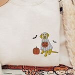 Sand crewneck sweatshirt laid flat beside a felt hat, embroidered with a yellow Labrador in a white ghost sheet with a pumpkin and two bats on the left chest