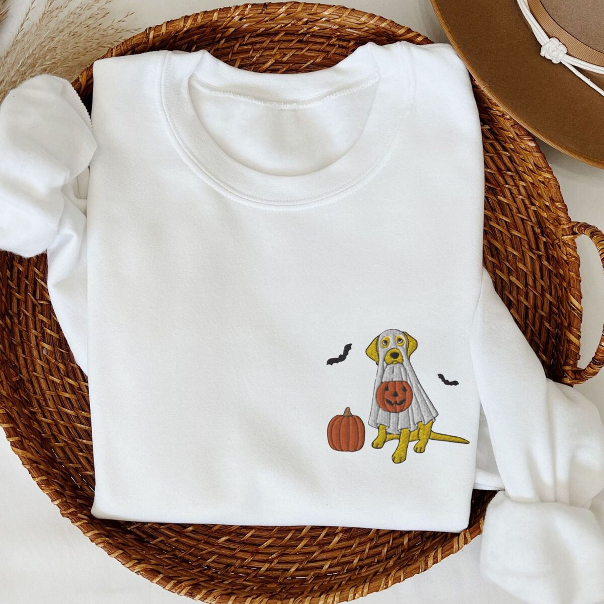 Yellow Lab Ghost Sweatshirt - White White crewneck sweatshirt folded in a wicker basket, embroidered with a yellow Labrador in a white ghost sheet holding a jack-o'-lantern bucket beside a pumpkin and bats
