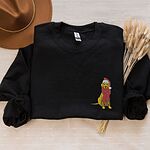 Black crewneck sweatshirt laid flat with a hat and dried pampas, embroidered with a small yellow Labrador in a red Santa hat and a long fringed red scarf on the left chest