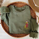 Military Green crewneck sweatshirt folded in a wicker basket with a hat, embroidered with a small yellow Labrador in a red Santa hat and a long fringed red scarf on the left chest