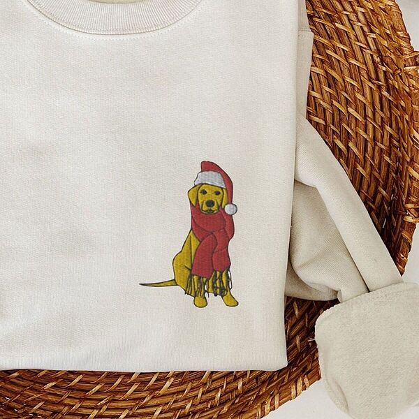 Sand crewneck sweatshirt folded in a wicker basket, embroidered with a small yellow Labrador in a red Santa hat and a long fringed red scarf on the left chest