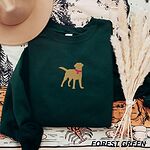 Forest Green crewneck sweatshirt laid flat with a hat and pampas grass, embroidered with a golden yellow Labrador silhouette standing in a pink collar and round tag in the center of the chest