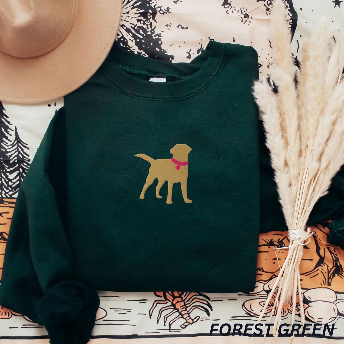 Yellow Lab Silhouette Sweatshirt – Forest Green Forest Green crewneck sweatshirt laid flat with a hat and pampas grass, embroidered with a golden yellow Labrador silhouette standing in a pink collar and round tag in the center of the chest
