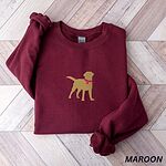 Maroon crewneck sweatshirt laid flat on herringbone wood, embroidered with a golden yellow Labrador silhouette standing in a pink collar and round tag in the center of the chest