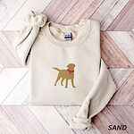 Sand crewneck sweatshirt laid flat on whitewashed wood, embroidered with a golden yellow Labrador silhouette standing in a pink collar and round tag in the center of the chest