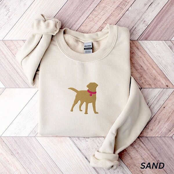 Sand crewneck sweatshirt laid flat on whitewashed wood, embroidered with a golden yellow Labrador silhouette standing in a pink collar and round tag in the center of the chest