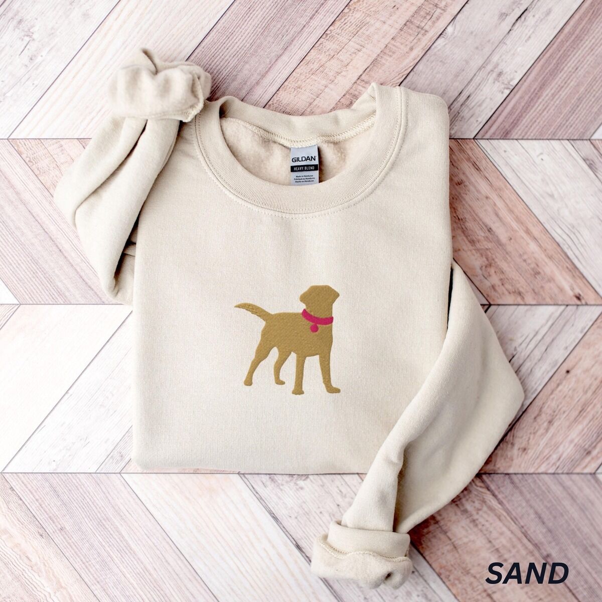 Yellow Lab Silhouette Sweatshirt – Sand Sand crewneck sweatshirt laid flat on whitewashed wood, embroidered with a golden yellow Labrador silhouette standing in a pink collar and round tag in the center of the chest
