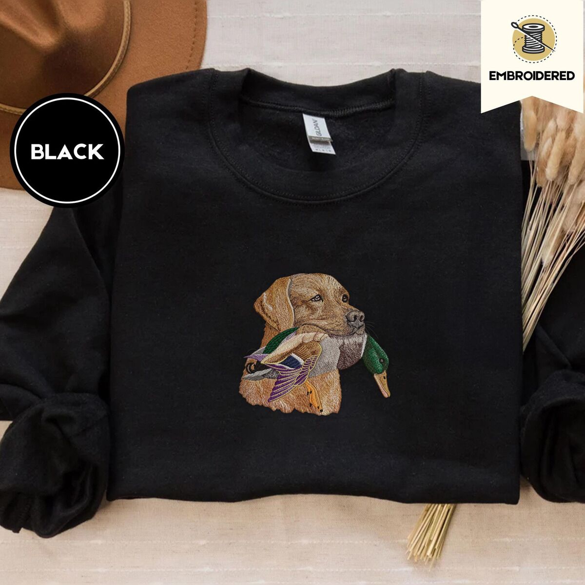 Yellow Lab Sweatshirt – Black Black crewneck sweatshirt with a brown felt hat and dried bunny tail grass, embroidered with a yellow Labrador head carrying a mallard drake in its mouth
