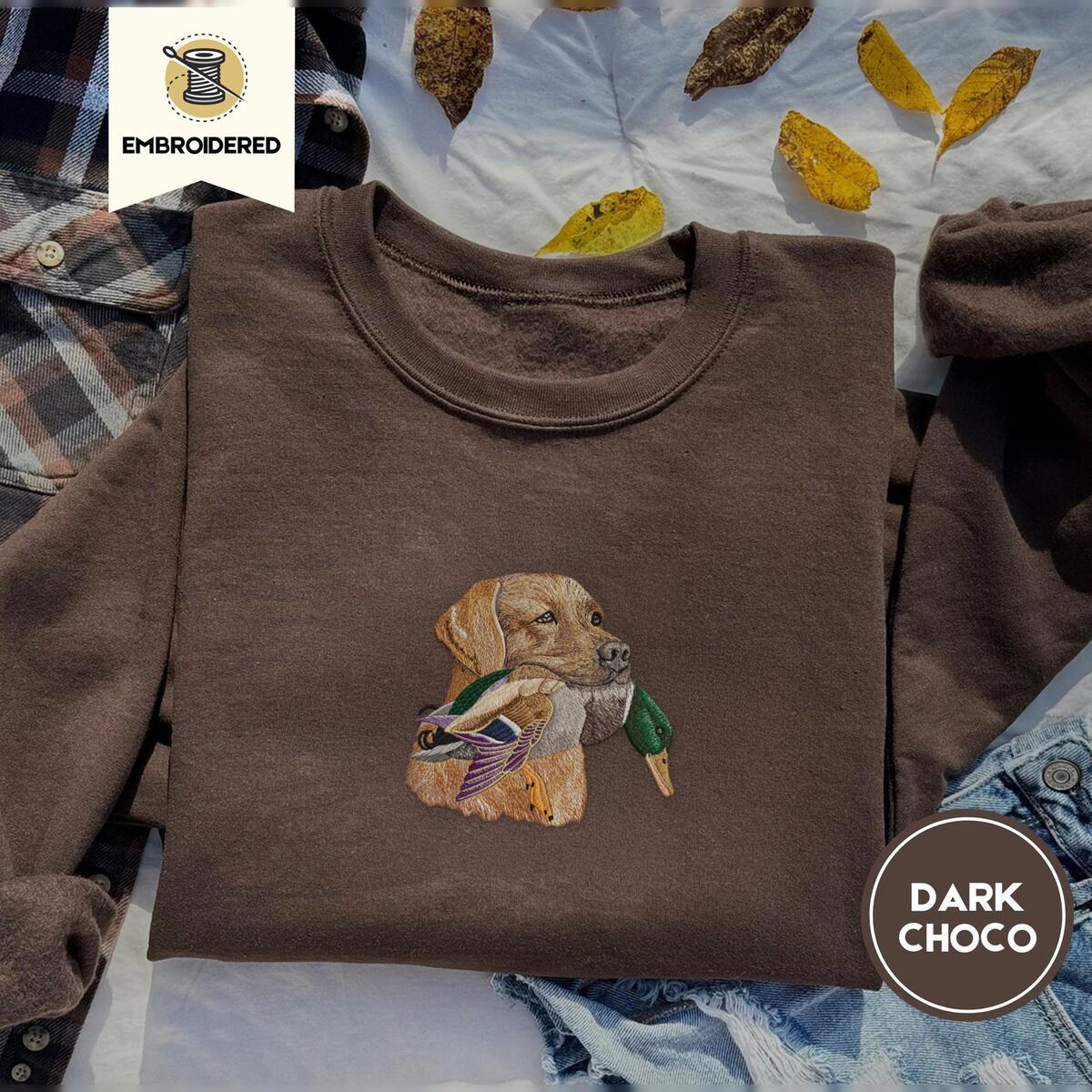 Yellow Lab Sweatshirt – Dark Chocolate Dark Chocolate crewneck sweatshirt on a plaid flannel shirt with yellow autumn leaves and denim shorts, embroidered with a yellow Labrador head carrying a mallard drake in its mouth