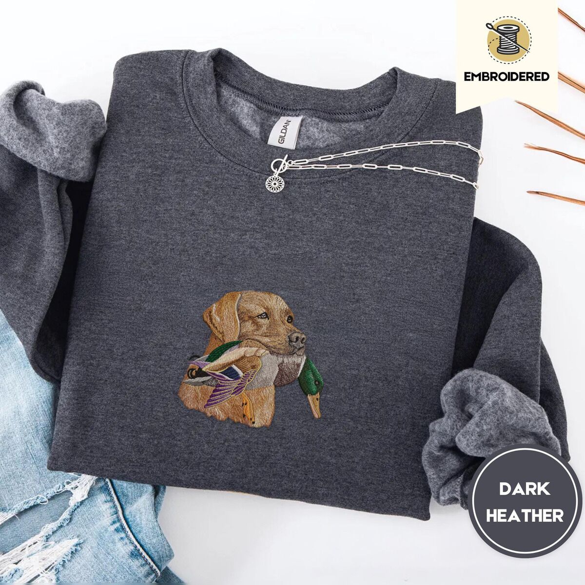 Yellow Lab Sweatshirt – Dark Heather Dark Heather crewneck sweatshirt folded with ripped jeans and a silver chain necklace, embroidered with a yellow Labrador head carrying a mallard drake in its mouth