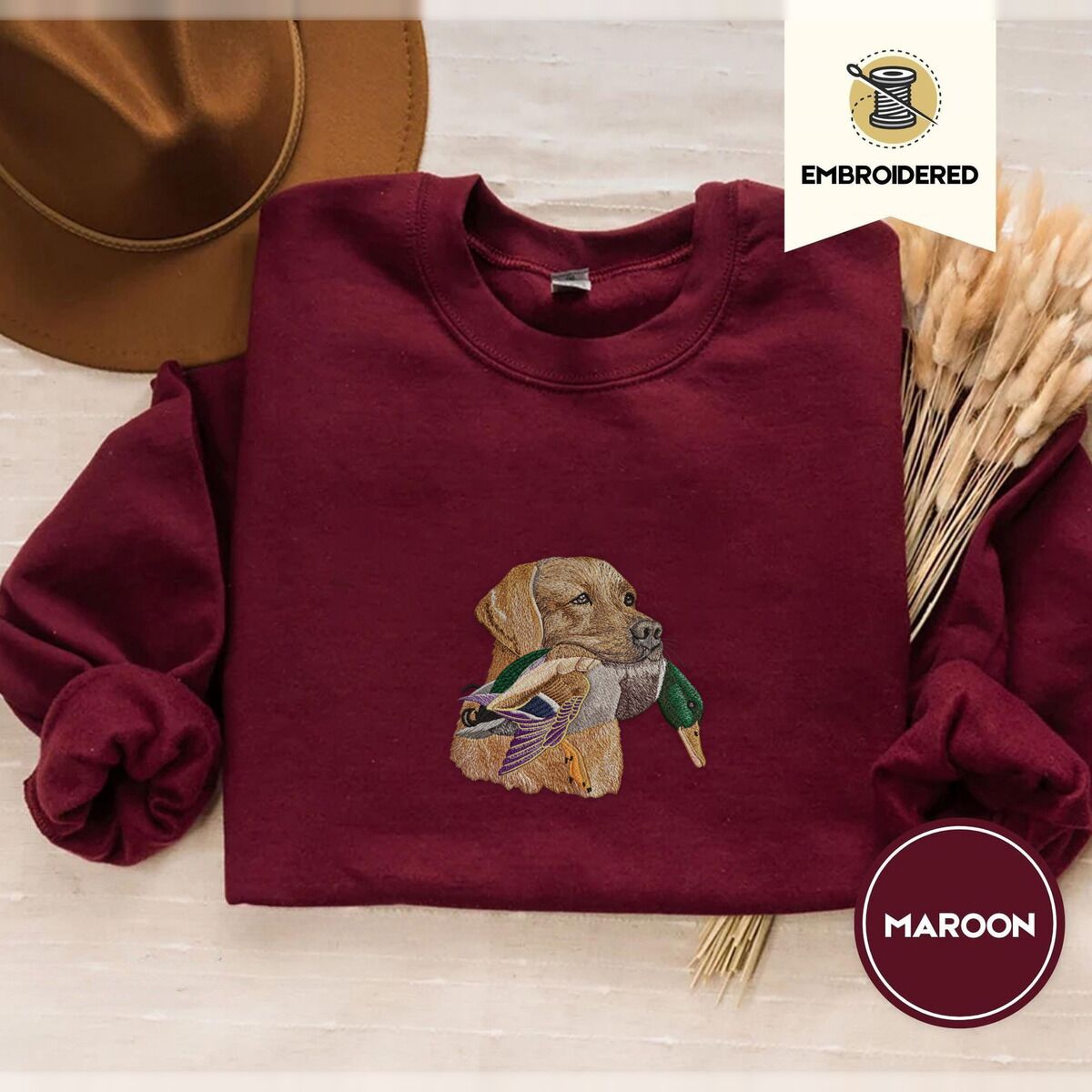 Yellow Lab Sweatshirt – Maroon Maroon crewneck sweatshirt with a brown felt hat and dried bunny tail grass, embroidered with a yellow Labrador head carrying a mallard drake in its mouth