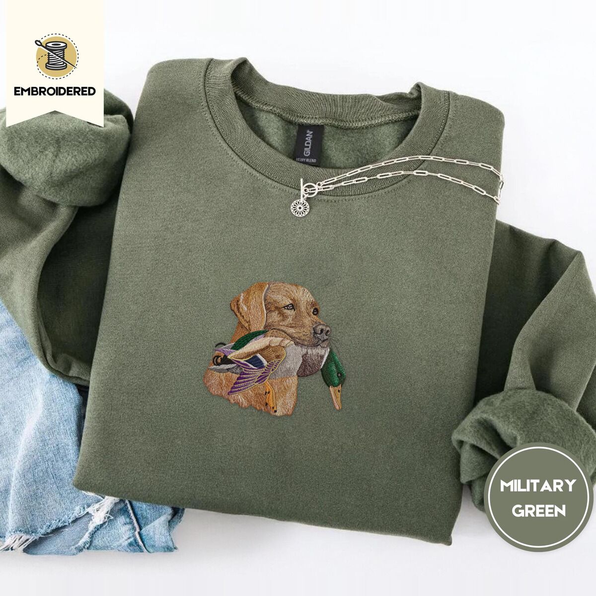 Yellow Lab Sweatshirt – Military Green Military Green crewneck sweatshirt folded with light jeans and a silver chain necklace, embroidered with a yellow Labrador head carrying a mallard drake in its mouth