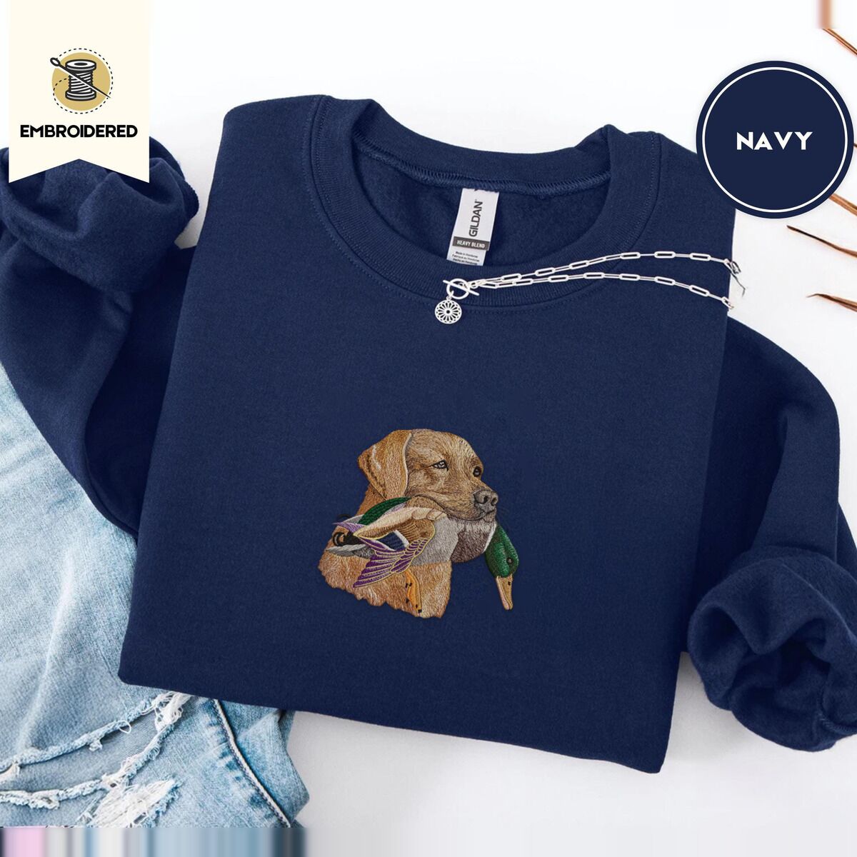 Yellow Lab Sweatshirt – Navy Navy crewneck sweatshirt folded with ripped jeans and a silver chain necklace, embroidered with a yellow Labrador head carrying a mallard drake in its mouth