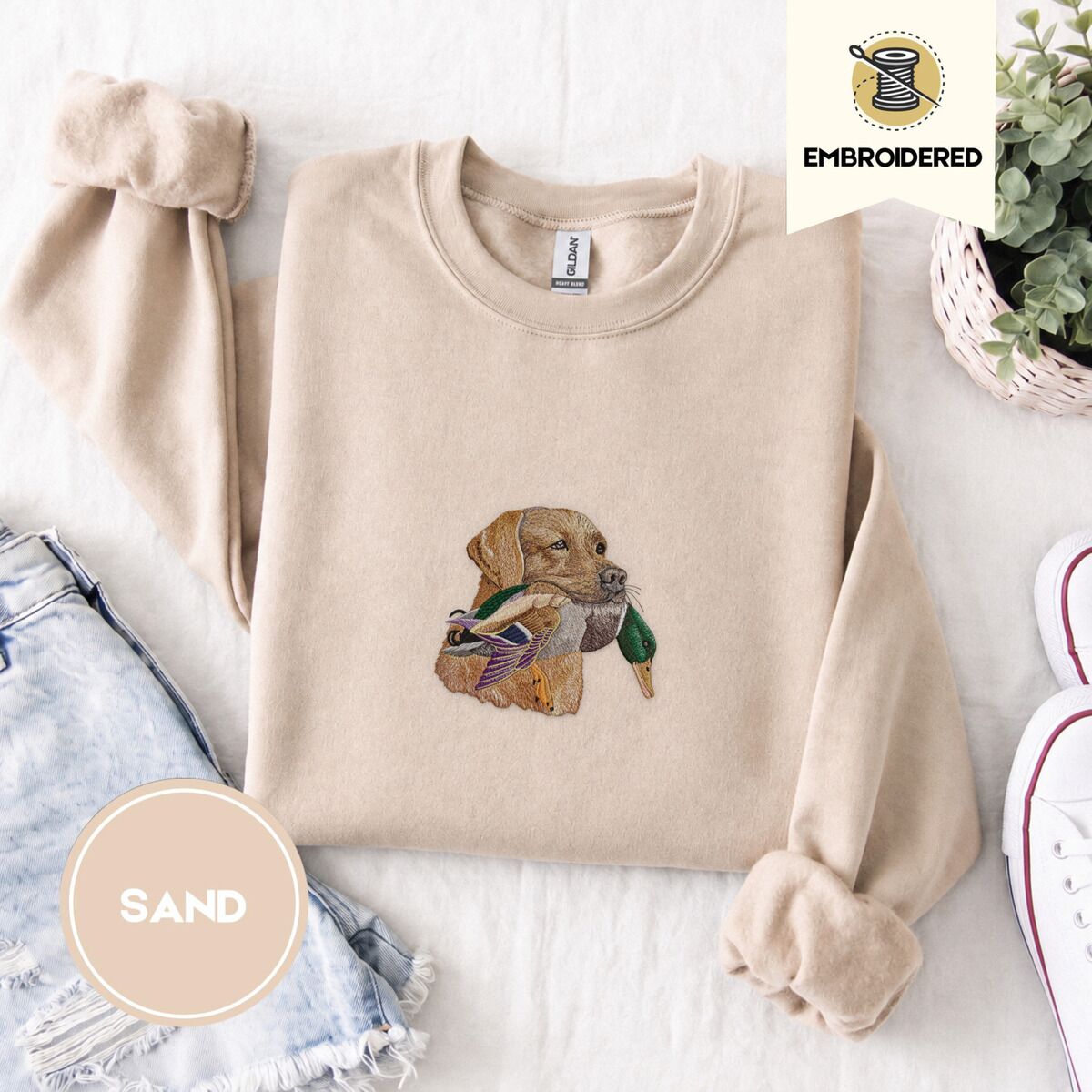 Yellow Lab Sweatshirt – Sand Sand crewneck sweatshirt folded with denim shorts, white sneakers and a potted plant, embroidered with a yellow Labrador head carrying a mallard drake in its mouth